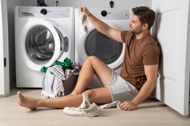 Çamaşır makinesinin yanında çamaşır sıralayan bir adam. Kirli çamaşırları olan bir çamaşır makinesinin yanında bir adam oturuyor. Çamaşır makinesinin yanında kirli bir çorap tutan bir adam, kirli ayakkabılar, kuru temizlemeci..