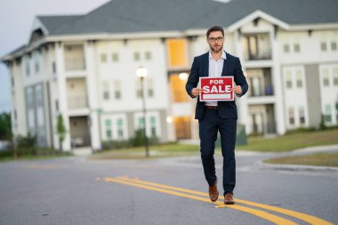 A man in a suit is holding a sign that says For Sale while walking down a street. real state agent in a suit walks down the street outside a new apartment complex with a new price offer clipart