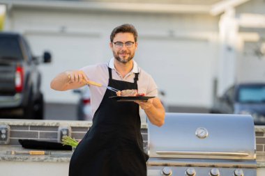 a man in an apron grilling a steak. Barbeque on holiday picnic. Man grilling a steak on BBQ. Grill and barbecue cook. Chef with BBQ cooking tools. Barbecue and grill. Picnic barbecue party. clipart