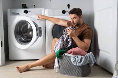 Çamaşır odasında çamaşır makinesinin yanında giysileri sıralayan mutlu bir adam. Adam kirli çamaşırlarıyla çamaşır makinesinin yanında bir evin zemininde oturuyor. Bir adam çamaşırlarını evde yıkıyor. Kirli çamaşırları olan bir adam.