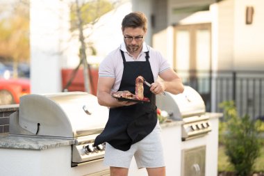 Önlüklü bir adam biftek pişiriyor. Tatil pikniğinde barbekü. Adam barbeküde biftek pişiriyor. Izgara ve barbekü aşçısı. Barbekü pişirme aletleri olan bir şef. Barbekü ve ızgara. Piknik barbekü partisi.