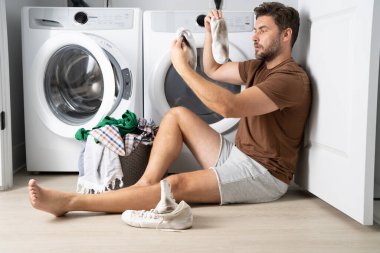 Çamaşır makinesinin yanında çamaşır sıralayan bir adam. Kirli çamaşırları olan bir çamaşır makinesinin yanında bir adam oturuyor. Çamaşır makinesinin yanında kirli bir çorap tutan bir adam, kirli ayakkabılar, kuru temizlemeci..