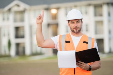 Miğfer ve inşaat yeleği içinde inşaat müfettişi. Yanında da pano ve tablet var. İnşaat alanında çalışan erkek işçi yeni bir binanın çizimlerine bakıyor.. 