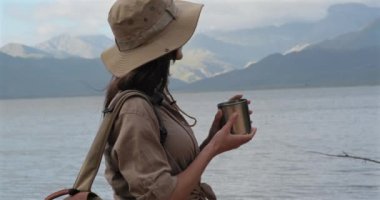 Sırt çantalı kadın yürüyüşçü dağlarda dinlenirken sıcak çay içiyor. Yürüyüşçü dağlardaki bir gölün yakınında sıcak bir içecek içer. Amerikalı bir turist Amerikan dağlarında tek başına yürüyüş yapıyor.