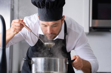 Profesyonel üniformalı modern şef. Kaşıkla tencereye çorba karıştıran erkek aşçının eline yakın plan. Yemek pişirme, aşçılık ve insan konsepti - erkek aşçı tencereli veya tencereli.