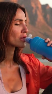 Female athlete replenishing water balance after fitness workout. Relaxed fit girl drink water from bottle. Close backlit portrait of active woman drinking from water bottle while resting from training