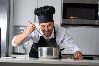 Profesyonel üniformalı modern şef. Kaşıkla tencereye çorba karıştıran erkek aşçının eline yakın plan. Yemek pişirme, aşçılık ve insan konsepti - erkek aşçı tencereli veya tencereli.