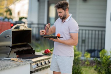 İspanyol bir adam ızgarada barbekü pişiriyor, önlük giyiyor. Erkek kavrulmuş biftek, ızgarada ızgara, arka bahçede ızgara, yaz aile pikniği, doğada yemek..