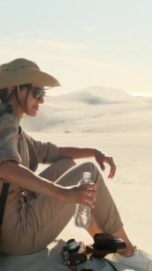 Young woman traveler looking sunset at red sand dunes. Female alone with a travel backpack in a desert background. stylish girl traveler in a hat and glasses drinks water in the desert.