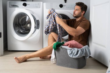 Bir adam banyonun zemininde oturuyor, kirli çamaşırları gri bir sepetten ayırıyor. Elbiselerinden birini çıkarmak için bir çamaşır makinesine uzanıyor. Banyo temiz ve modern. 