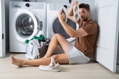 Çamaşır makinesinin yanında çamaşır sıralayan bir adam. Kirli çamaşırları olan bir çamaşır makinesinin yanında bir adam oturuyor. Çamaşır makinesinin yanında kirli bir çorap tutan bir adam, kirli ayakkabılar, kuru temizlemeci..