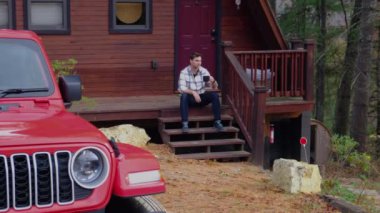 hippi adam ahşap evin verandasında oturuyor ve off-road yolculuğundan sonra aracına bakıyor. Olgun bir adam dağ evinin verandasında sıcak bir içecekten hoşlanır. Karlı kış doğasında tatilde olan olgun bir adam..