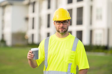 İnşaat işçisi inşaat alanında kahve molası veriyor. Miğferli ve gözlüklü bir adam sıcak bir içecek içer. Olgun inşaat işçisi mola verip kahve içiyor.