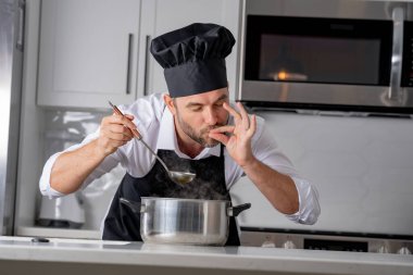 Profesyonel üniformalı modern şef. Kaşıkla tencereye çorba karıştıran erkek aşçının eline yakın plan. Yemek pişirme, aşçılık ve insan konsepti - erkek aşçı tencereli veya tencereli.