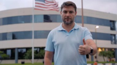 Başarılı bir yetişkin erkek, Amerikan bayrağının yanında kendine güvenen bir baş parmak gösteriyor. Başparmakları havada beyaz bir adam, çünkü iyi bir şey oldu. Başarı için mutlu ve gülümseyen parmaklar. Kazanan jest.