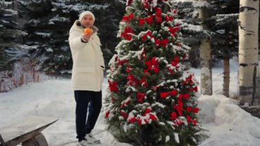Dünyevi ceketli ve şapkalı bir adam Noel ağacının yanında elinde oyuncaklarla elinde portakal tutuyordu. Kışın kar altında gülümseyen ve kameraya mandalina gösteren bir adam. Bir adam Noel ağacı süslüyor.
