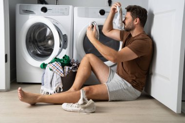 Çamaşır makinesinin yanında çamaşır sıralayan bir adam. Kirli çamaşırları olan bir çamaşır makinesinin yanında bir adam oturuyor. Çamaşır makinesinin yanında kirli bir çorap tutan bir adam, kirli ayakkabılar, kuru temizlemeci..