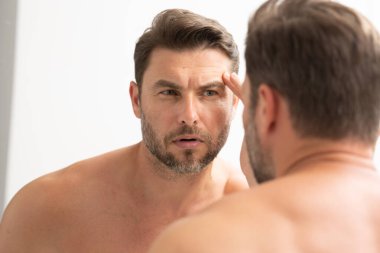 Bakımlı, yetişkin bir adam aynada düzgün, kırışık olmayan bir cilde bakar. Yakışıklı adam aynada kırışıklıkları kontrol ediyor. Aynanın önündeki adam kırışıklıklarına bakıyor.