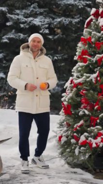 Dünyevi ceketli ve şapkalı bir adam Noel ağacının yanında elinde oyuncaklarla elinde portakal tutuyordu. Kışın kar altında gülümseyen ve kameraya mandalina gösteren bir adam. Bir adam Noel ağacı süslüyor.