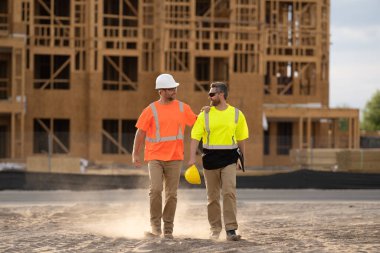İnşaat mühendisleri inşaat alanında, bitmemiş bir evin yanında tozun içinden yürüyorlar. Açık hava inşaat sahasında çalışan mühendis ve işçi planı