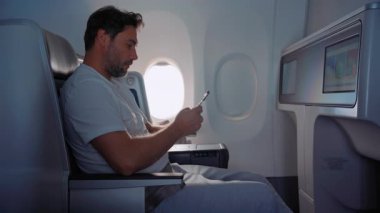 Elinde telefon olan mutlu bir adam uçakta telefonunun üzerinde oturuyor. Uçakla seyahat ederken telefonu kullanan bir adam. Mutlu adam telefon kullanır ve uçuş sırasında uçakta kulaklık dinler..