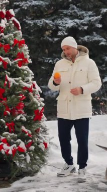 Dünyevi ceketli ve şapkalı bir adam Noel ağacının yanında elinde oyuncaklarla elinde portakal tutuyordu. Kışın kar altında gülümseyen ve kameraya mandalina gösteren bir adam. Bir adam Noel ağacı süslüyor.
