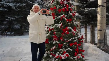 Noel ağacının yanında kışlık ceketli bir adam elleriyle bir kalp gösteriyor. Adam Aşkı Göstermek İçin Elleriyle Kalp Şekli Yapıyor. Adam elleriyle kalp sembolü yapıyor. Bir adam ve Noel neşesi