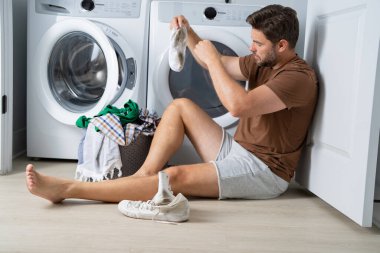 Bir adam banyonun zemininde oturuyor, kirli çamaşırları gri bir sepetten ayırıyor. Elbiselerinden birini çıkarmak için bir çamaşır makinesine uzanıyor. Banyo temiz ve modern. 