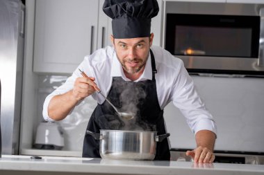 Profesyonel üniformalı modern şef. Kaşıkla tencereye çorba karıştıran erkek aşçının eline yakın plan. Yemek pişirme, aşçılık ve insan konsepti - erkek aşçı tencereli veya tencereli.
