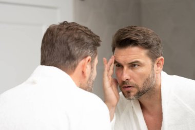 Bakımlı, yetişkin bir adam aynada düzgün, kırışık olmayan bir cilde bakar. Yakışıklı adam aynada kırışıklıkları kontrol ediyor. Aynanın önündeki adam kırışıklıklarına bakıyor.