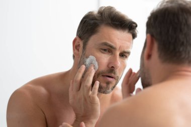 Banyoda yüz kremi süren yakışıklı bir adam. Yüz kreminden sonra tatmin olmuş genç bir adamın yakından güzel portresi. Çekici esmer adam tenine dokunuyor. Erkek kozmetik ve güzellik konsepti