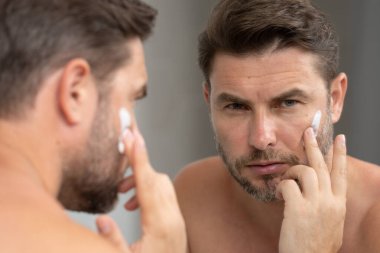 Banyoda yüz kremi süren yakışıklı bir adam. Yüz kreminden sonra tatmin olmuş genç bir adamın yakından güzel portresi. Çekici esmer adam tenine dokunuyor. Erkek kozmetik ve güzellik konsepti