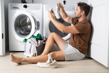 Bir adam banyonun zemininde oturuyor, kirli çamaşırları gri bir sepetten ayırıyor. Elbiselerinden birini çıkarmak için bir çamaşır makinesine uzanıyor. Banyo temiz ve modern. 