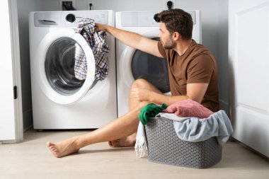 Bir adam banyonun zemininde oturuyor, kirli çamaşırları gri bir sepetten ayırıyor. Elbiselerinden birini çıkarmak için bir çamaşır makinesine uzanıyor. Banyo temiz ve modern. 