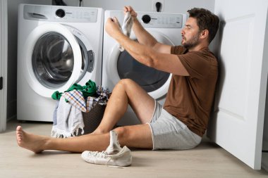 Çamaşır odasında çamaşır makinesinin yanında giysileri sıralayan mutlu bir adam. Adam kirli çamaşırlarıyla çamaşır makinesinin yanında bir evin zemininde oturuyor. Bir adam çamaşırlarını evde yıkıyor. Kirli çamaşırları olan bir adam.