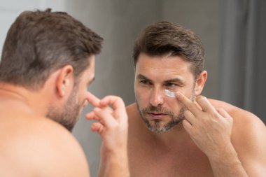 Banyoda yüz kremi süren yakışıklı bir adam. Yüz kreminden sonra tatmin olmuş genç bir adamın yakından güzel portresi. Çekici esmer adam tenine dokunuyor. Erkek kozmetik ve güzellik konsepti