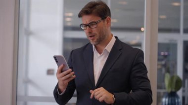 Resmi giyinmiş iş adamı, telefonundan üzücü haberler okurken göğsünü sıkıca tutuyor. Duygu, modern bir ofis ortamında kurumsal gerilim, siyasi etki veya ani yatırım kaybı gösteriyor.