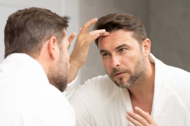 Orta yaşlı bir adam banyo aynasında kırışıklıkları ve deri dokusunu dikkatlice inceler. Erkek güzelliği ve yaşlanma konsepti kozmetik kaygıları vurguluyor. Sahne tımar, cilt bakımı ve estetik bakımı yansıtır..