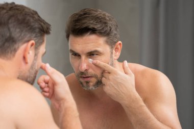 Banyoda yüz kremi süren yakışıklı bir adam. Yüz kreminden sonra tatmin olmuş genç bir adamın yakından güzel portresi. Çekici esmer adam tenine dokunuyor. Erkek kozmetik ve güzellik konsepti