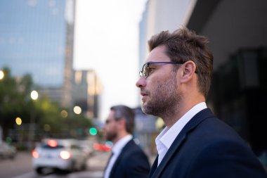 İki işadamı, yoğun bir şehir caddesinde dikiliyor. Odaklanma ve kararlılıkla bekliyorlar. Sahne kentsel yaşam tarzını, liderlik anlayışını, fırsatları ve profesyonel hırsı yansıtıyor.