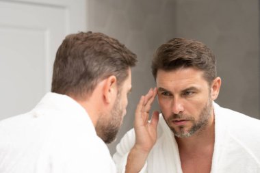 Bakımlı, yetişkin bir adam aynada düzgün, kırışık olmayan bir cilde bakar. Yakışıklı adam aynada kırışıklıkları kontrol ediyor. Aynanın önündeki adam kırışıklıklarına bakıyor.