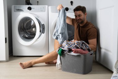 Çamaşır odasında çamaşır makinesinin yanında giysileri sıralayan mutlu bir adam. Adam kirli çamaşırlarıyla çamaşır makinesinin yanında bir evin zemininde oturuyor. Bir adam çamaşırlarını evde yıkıyor. Kirli çamaşırları olan bir adam.