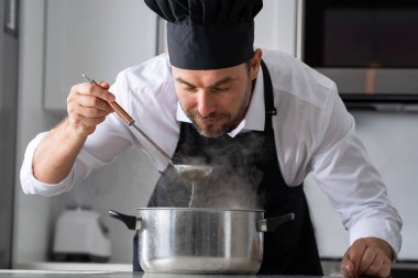 Profesyonel üniformalı modern şef. Kaşıkla tencereye çorba karıştıran erkek aşçının eline yakın plan. Yemek pişirme, aşçılık ve insan konsepti - erkek aşçı tencereli veya tencereli.