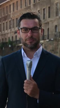 Muhabir, bir şehir caddesinde el mikrofonuyla canlı bir güncelleme sunuyor. Erkek gazeteci güncel olayları takip ediyor ve hızlı röportajlar yapıyor. Sahne medya çalışmaları, aciliyet ve şehir haberlerini gösteriyor.