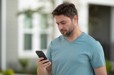 Gündelik giysiler içinde genç bir adam akıllı telefon kullanırken dışarıda duruyor. Erkek, sohbet etmek için mobil uygulamaları, sosyal ağları, hatırlatıcıları ve günlük görev organizasyonlarını kontrol eder. Adam akıllı telefon uygulamalarını kontrol ediyor.