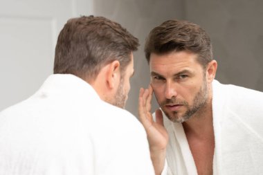 Bakımlı, yetişkin bir adam aynada düzgün, kırışık olmayan bir cilde bakar. Yakışıklı adam aynada kırışıklıkları kontrol ediyor. Aynanın önündeki adam kırışıklıklarına bakıyor.