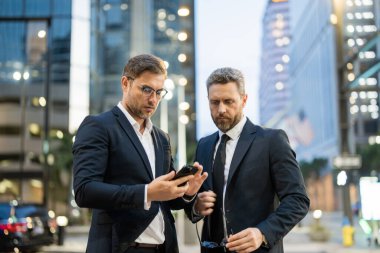 Girişimciler modern bir şehirde dururken akıllı telefondaki bilgileri analiz ederler. Resmi takım elbiseli iki erkek işadamı odaklanma ve konsantrasyonla stratejiyi tartışıyor. Sahne hırs ve takım çalışmasını gösteriyor