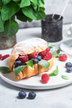 Beyaz tabakta ahududu, yaban mersini ve krem peynirli kruvasan.