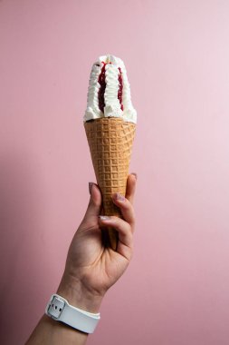 Bir kadının eli lezzetli bir dondurma külahını tutuyor. Krem şantili ve çilek soslu.