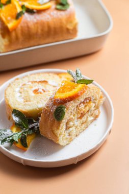 Parlak bir arka planda beyaz bir tabakta kremalı ve naneli turuncu sünger rulosu.